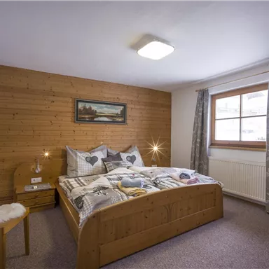 A cozy bedroom with a wooden bed and a warm, inviting atmosphere. The room is simply furnished, with a chair and a window that lets in daylight.