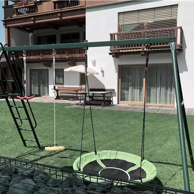 Ein Spielplatz mit einer Schaukel und einem Wippbrett in einem Garten. Im Hintergrund sind ein Haus und eine Terrasse mit Fenster zu sehen.