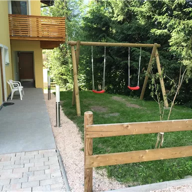 Ein Spielplatz mit einer Schaukel steht neben einem gelben Gebäude. Der Bereich ist von einem Holzzaun umgeben und von Bäumen umgeben.