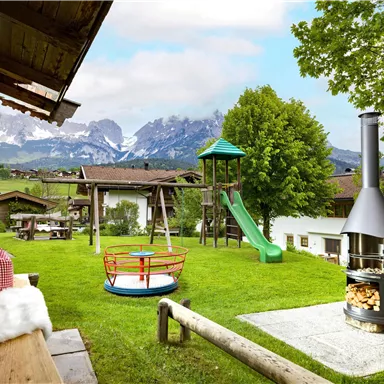 A beautiful garden with a playground and slide. In the background, the mountains and a traditional wooden house can be seen.