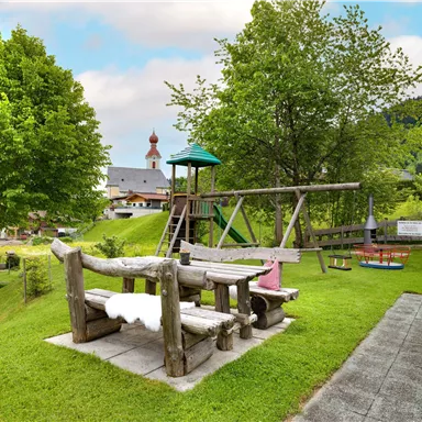 A playground with a swing and slide is located in a green meadow. In the background, trees and a building are visible.