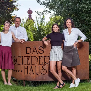 A group of four people stands in front of a rustic sign that reads "DAS SCHEDER HAUS". In the background, green plants and a beautiful garden can be seen.