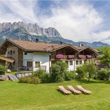 A tranquil chalet in the greenery with blooming flowers. In the background, the mountains rise majestically under a clear sky.