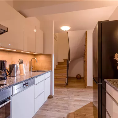 A modern kitchen with white cabinets and an elegant countertop. In the background, a staircase leads to the upper rooms.