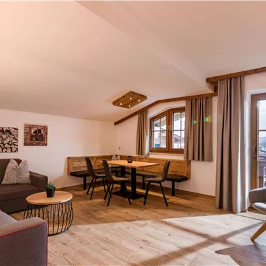 A spacious living room with hardwood flooring and large windows. There is a sofa, a dining table, and decorative wall art.