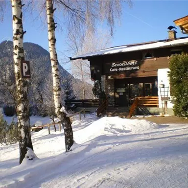 A snow-covered restaurant with a rustic wooden facade. In the background, snow-capped mountains and a bright blue sky can be seen.