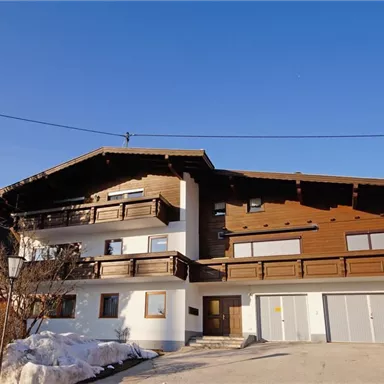 A modern wooden house in the mountains, surrounded by snow-covered trees. The clear blue sky complements the alpine atmosphere.