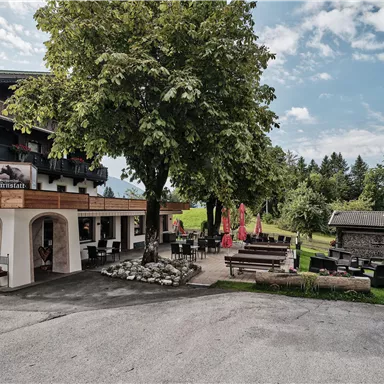 A cozy inn in a green environment. In front of the building, there are seating areas and a large tree.