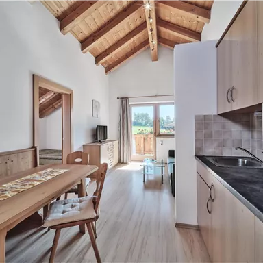A bright, cozy living room with a wooden ceiling and an open kitchen. Large windows provide plenty of daylight and a view outside.
