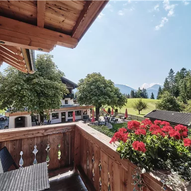 A cozy balcony view with red flowers and a glimpse of a green landscape. In the background, mountains and a blue sky are visible.