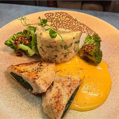 A beautifully arranged plate with stuffed chicken, creamy rice, and broccoli. It is accompanied by an orange sauce and a crispy decoration.