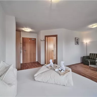 A bright, modern bedroom with a bed and a sofa. The walls are in neutral colors, and there is a friendly atmosphere.