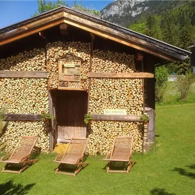 A rustic wooden house covered with stacked firewood. On top, there are two sun loungers on a green meadow.