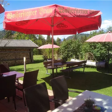 An inviting garden with tables and chairs under red sun umbrellas. In the background, green trees and a wooden structure are visible.