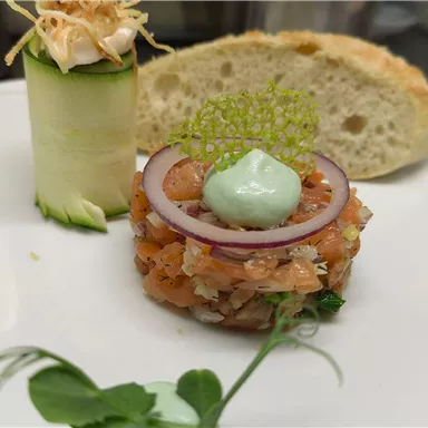 An artistically arranged plate with a salmon tartare tower, garnished with onion rings and a green sauce. Next to it lies a piece of fresh bread and a zucchini roll.