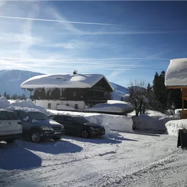 A snow-covered landscape with a traditional chalet and mountains in the background. The sky is clear and blue.