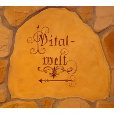A decorative stone with the inscription "Vital-world". The stone is embedded in a rustic wall made of rocks.