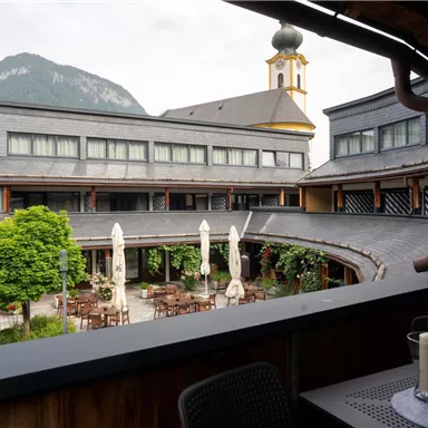 A courtyard with tables and chairs, surrounded by plants. In the background, buildings with a church tower and mountains can be seen.