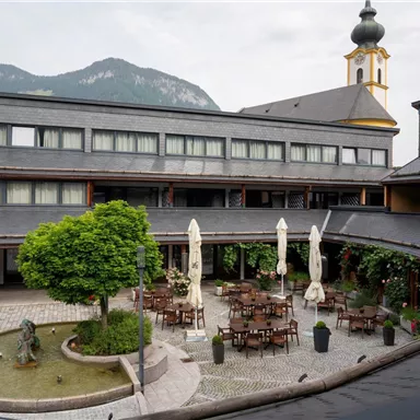 A modern-designed courtyard with tables and chairs. In the background, mountains and a church tower are visible.