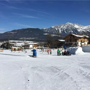 Eine schneebedeckte Landschaft mit Bergen im Hintergrund. Im Vordergrund sind Menschen beim Skifahren und Spielen im Schnee.