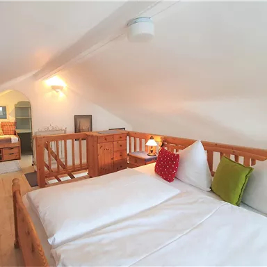 Ein gemütliches Dachzimmer mit einem großen Bett und Holzmöbeln. Es gibt eine warme Beleuchtung und farbige Kissen für zusätzlichen Komfort.