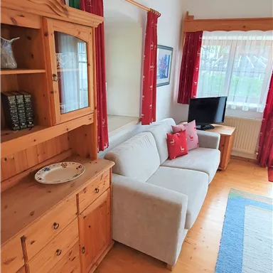 Ein gemütliches Wohnzimmer mit einem grauen Sofa und roten Kissen. Der Raum ist hell und hat eine Holzkommode sowie einen Fernseher.