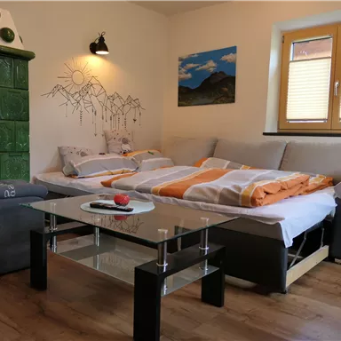 Ein gemütlicher Wohnraum mit einem Schlafsofa und einem Glastisch. An der Wand hängt ein Bild einer Berglandschaft, und ein grüner Kachelofen ist sichtbar.