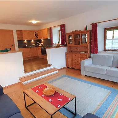 Ein gemütliches Wohnzimmer mit Holzfußboden und einer modernen Küche. Die Möbel sind schlicht und einladend, mit roten Vorhängen und einem Teppich in sanften Farben.