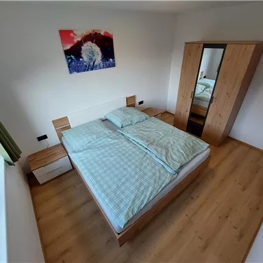 Ein gemütliches Schlafzimmer mit einem Doppelbett und einem großen Spiegel. An der Wand hängt ein buntes Bild, und der Boden ist aus hellem Holz.