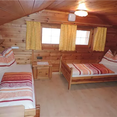 Ein gemütliches Zimmer mit Holzwänden und zwei Betten. Die Fenster sind mit hellen Vorhängen dekoriert und es gibt eine warmes Licht von der Deckenlampe.