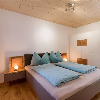 A cozy bedroom with a double bed and blue pillows. The walls are light and there are windows with orange curtains.