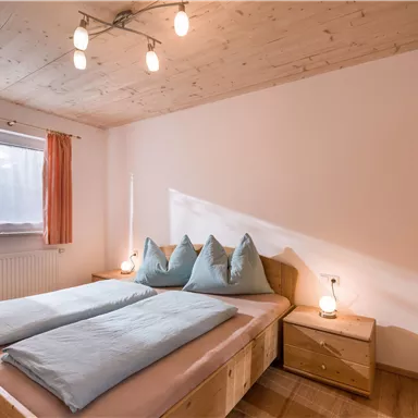 A cozy bedroom with a double bed and blue pillows. The walls are light, and the room has a window with curtains.