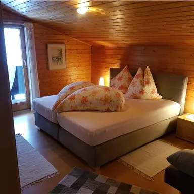 A cozy bedroom with wooden paneling and a double bed. There are soft pillows and a balcony with a view.