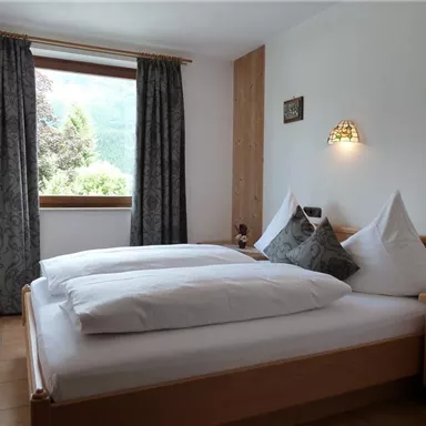 Ein gemütliches Schlafzimmer mit einem Doppelbett und zwei Kopfkissen. Große Fenster dekoriert mit Vorhängen bieten Blick auf die Natur draußen.