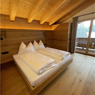 Ein gemütliches Schlafzimmer mit einem großen Doppelbett und Holzvertäfelung. Helle Wände und ein Balkonfenster sorgen für eine einladende Atmosphäre.
