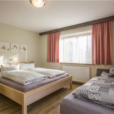 A modern bedroom with a double bed and a single bed. Bright windows and cozy decor create an inviting atmosphere.