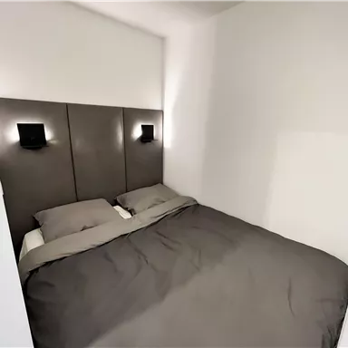 Ein modernes Schlafzimmer mit einem großen Bett und schlichten grauen Bettwaren. Die Wände sind in einem neutralen Weiß gehalten und es gibt stilvolle Wandlampen.