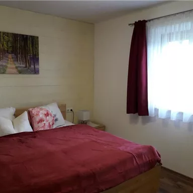 Ein gemütliches Schlafzimmer mit einem großen Bett und roten Kissen. Das Fenster lässt viel Sonnenlicht herein und ein schönes Bild von einem Wald hängt an der Wand.