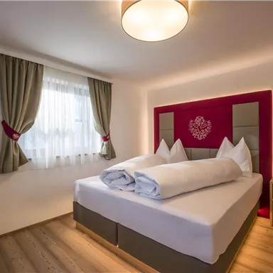 Ein modernes Schlafzimmer mit einem Doppelbett und zwei Kopfkissen. An der Wand hängt ein Fernseher, und die Fenster sind mit Vorhängen geschmückt.