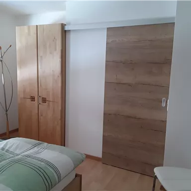 A modern bedroom with a wooden wardrobe and a sliding door. The walls are light, and there is a bed with striped bedding.
