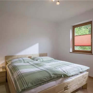 A simple bedroom with a double bed and checkered bedspreads. The windows are fitted with red roller blinds and let light in.