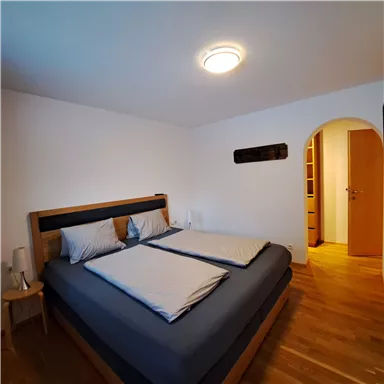 Ein helles Schlafzimmer mit einem großen Bett und Holzfußboden. Die Wände sind weiß, und es gibt einen offenen Zugang zu einem weiteren Raum.