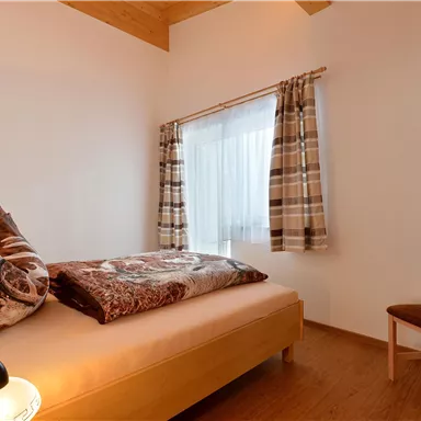 Ein gemütliches Schlafzimmer mit einem einzelnen Bett und einem Stuhl. Helle Wände und ein Fenster mit Vorhängen sorgen für eine freundliche Atmosphäre.