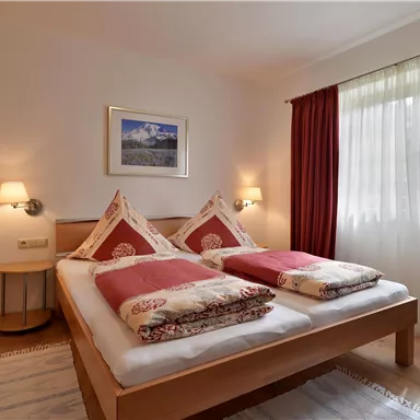 A cozy bedroom with two beds and red cushions. Large windows provide plenty of daylight and a pleasant atmosphere.
