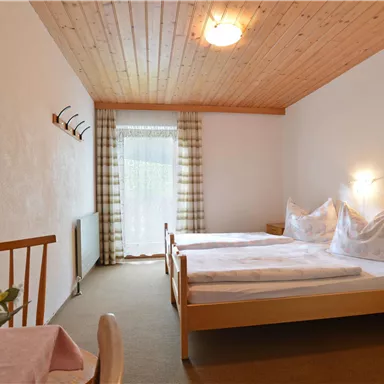 Ein einfaches Schlafzimmer mit zwei betten. Es gibt einen Tisch mit einer Blumenvase und großen Fenstern, die viel Licht hereinlassen.