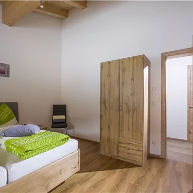 Ein modernes Schlafzimmer mit einem großen Bett und grünen Kissen. Im Hintergrund steht ein Holzschrank und eine offene Tür führt in einen anderen Raum.