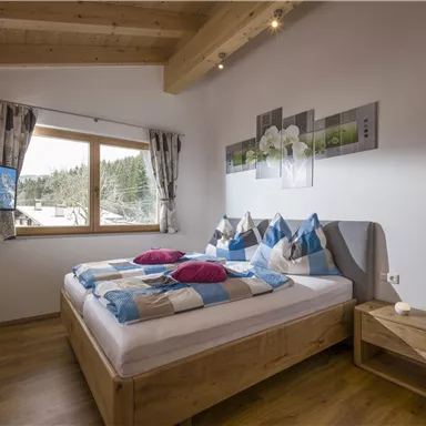 Ein gemütliches Schlafzimmer mit einem großen Fenster und Aussicht auf die Berge. Das Bett ist mit bunten Kissen dekoriert und es gibt einen Fernseher im Raum.