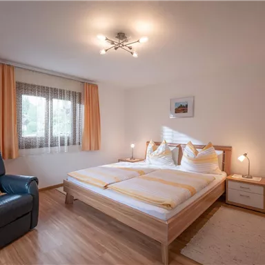 Ein gemütliches Schlafzimmer mit einem großen Bett und hellen Vorhängen. Es gibt einen bequemen Sessel und eine moderne Lampe.