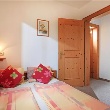 Ein gemütliches Schlafzimmer mit zwei Betten und bunten Kissen. Die Zimmerwände sind hell, und eine Tür führt zu einem weiteren Raum.