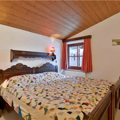 Ein gemütliches Schlafzimmer mit einem großen Bett und buntem Bettzeug. An der Wand befinden sich Holzverkleidungen und ein Fenster mit Vorhängen.